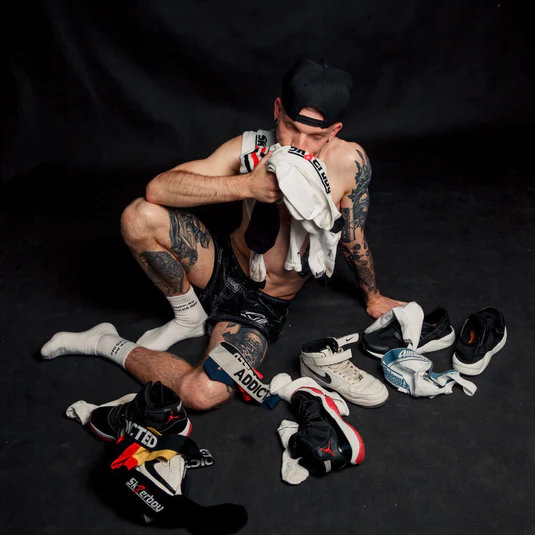 Vor schwarzem Hintergrund sitzt ein Sk8erboy inmitten von vielen Sneakern, Socks und Underwaer und snifft an mehreren white socks. Für die Produkte kann ein Gutschein beim Sk8erboy® Gewinnspiel auf der SUBVERSIVE gewonnen werden